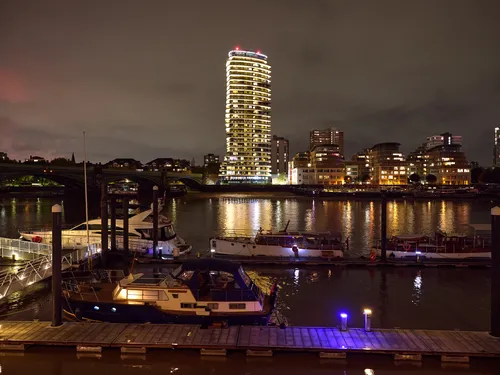 London view accross the thames from Imperial Wharf at night This urban night-time photograph captures a view across the River Thames in London from Imperial Wharf, taken in early autumn. The image highlights the illuminated riverside buildings and residential towers, reflecting brightly on the water, creating a vibrant atmosphere characteristic of the United Kingdom’s cityscape. Boats are moored in the foreground along the riverbank, contributing to the dynamic river scene. The architecture stands out against the night sky, demonstrating London’s modern riverside development and the lively ambiance along this stretch of the River Thames.